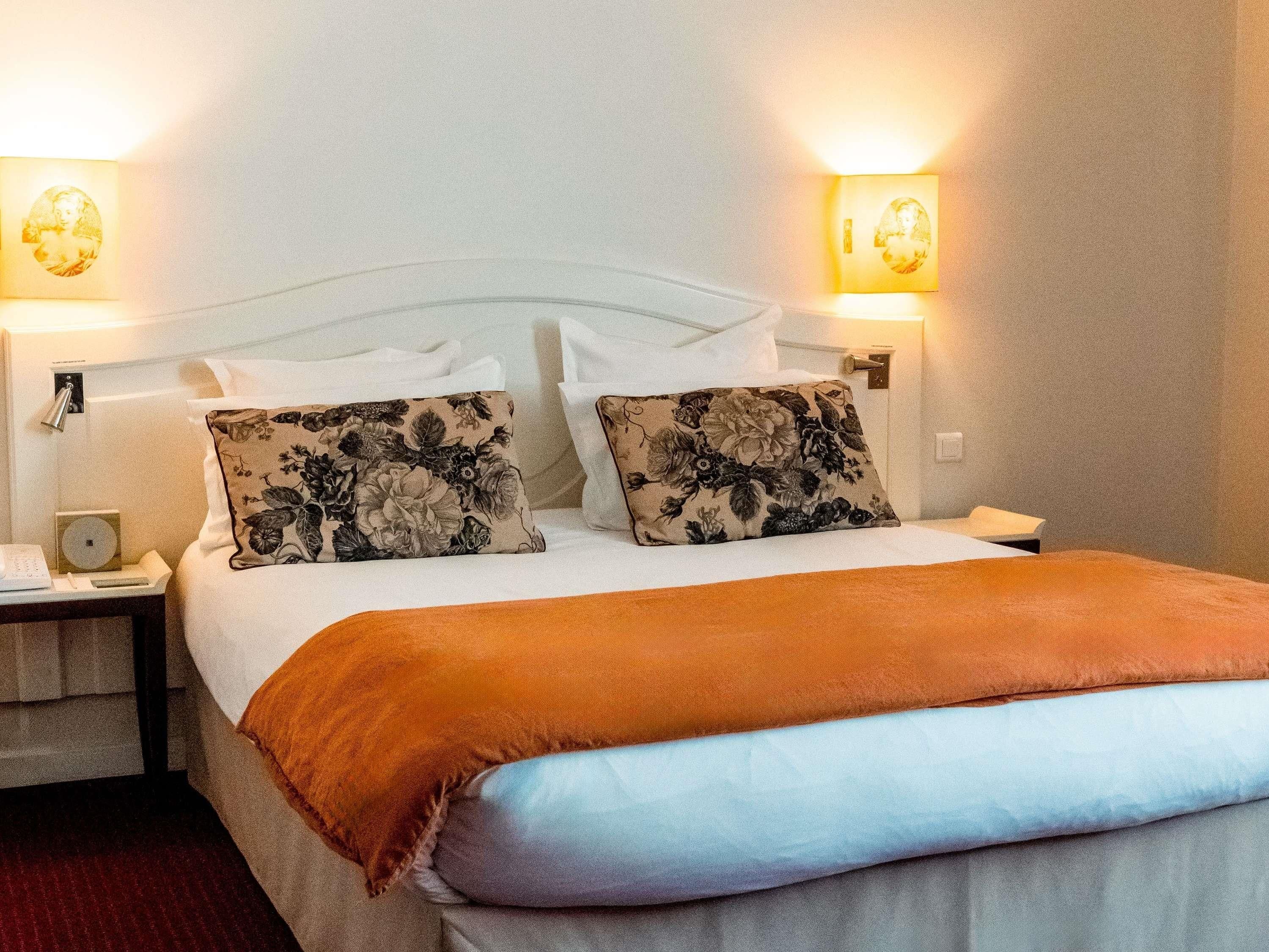 A hotel room with a bed, two pillows, a nightstand, and a wall-mounted lamp.