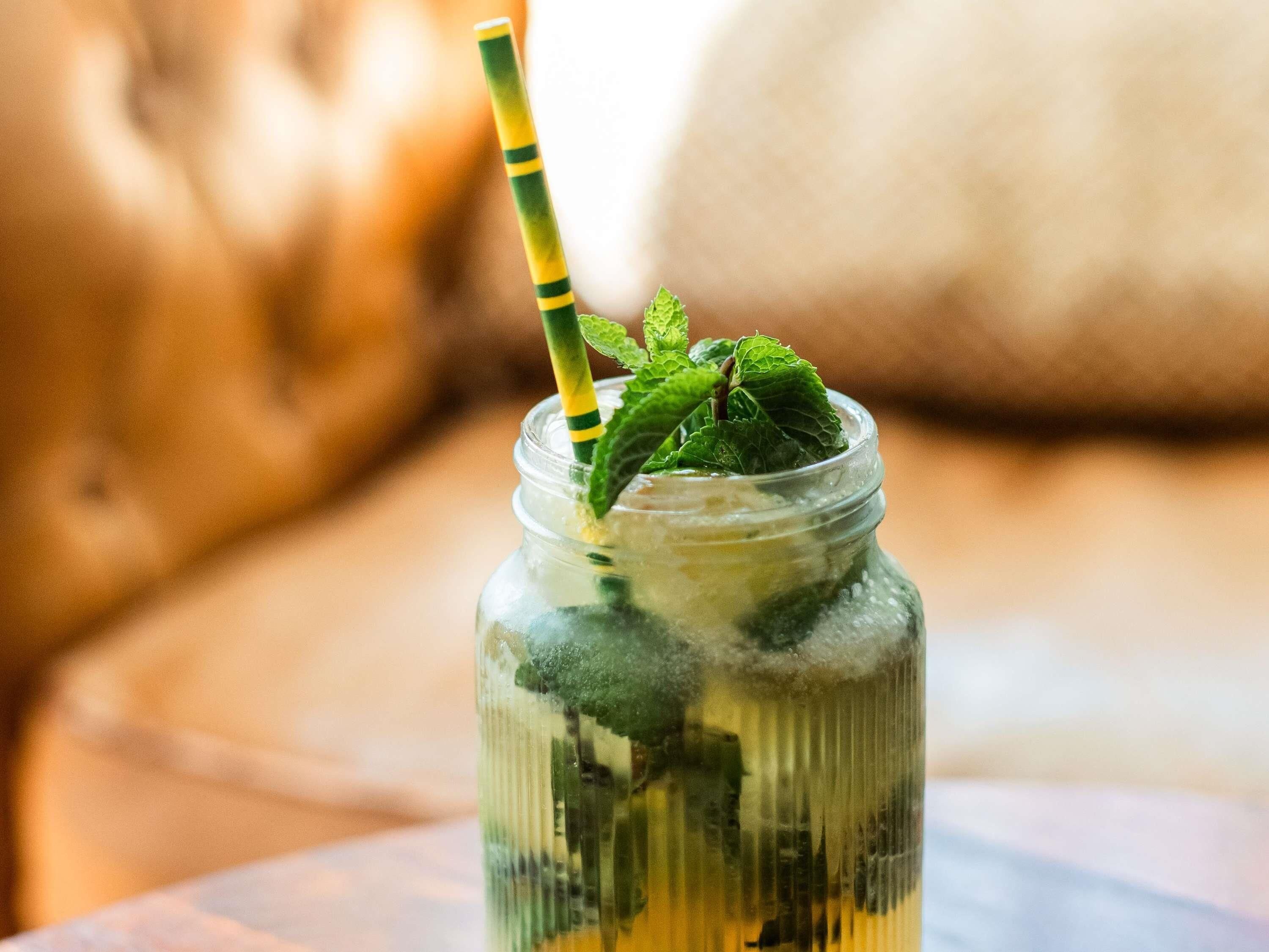 A clear glass jar with a green and yellow striped straw, filled with a beverage and mint leaves, placed on a surface.