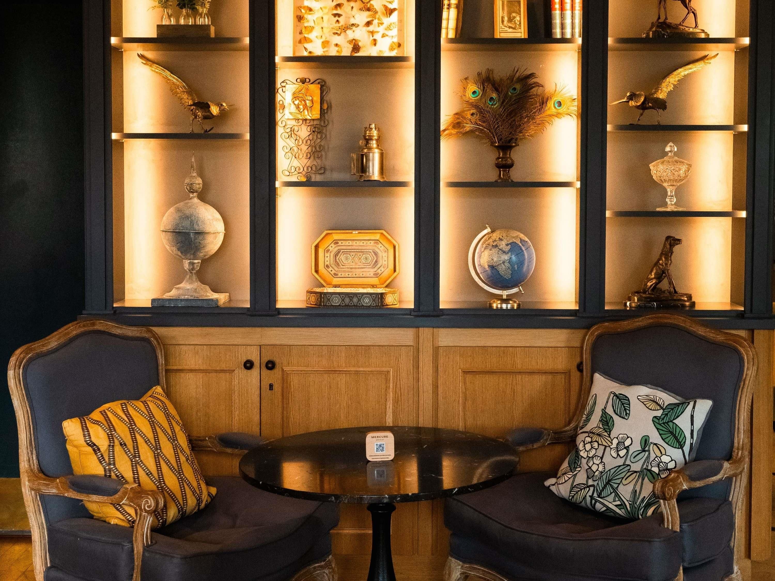 Een kamer met een houten kastje met diverse decoratieve objecten en een wereldbol. Er staan twee stoelen met kussens en een kleine ronde tafel in het midden.