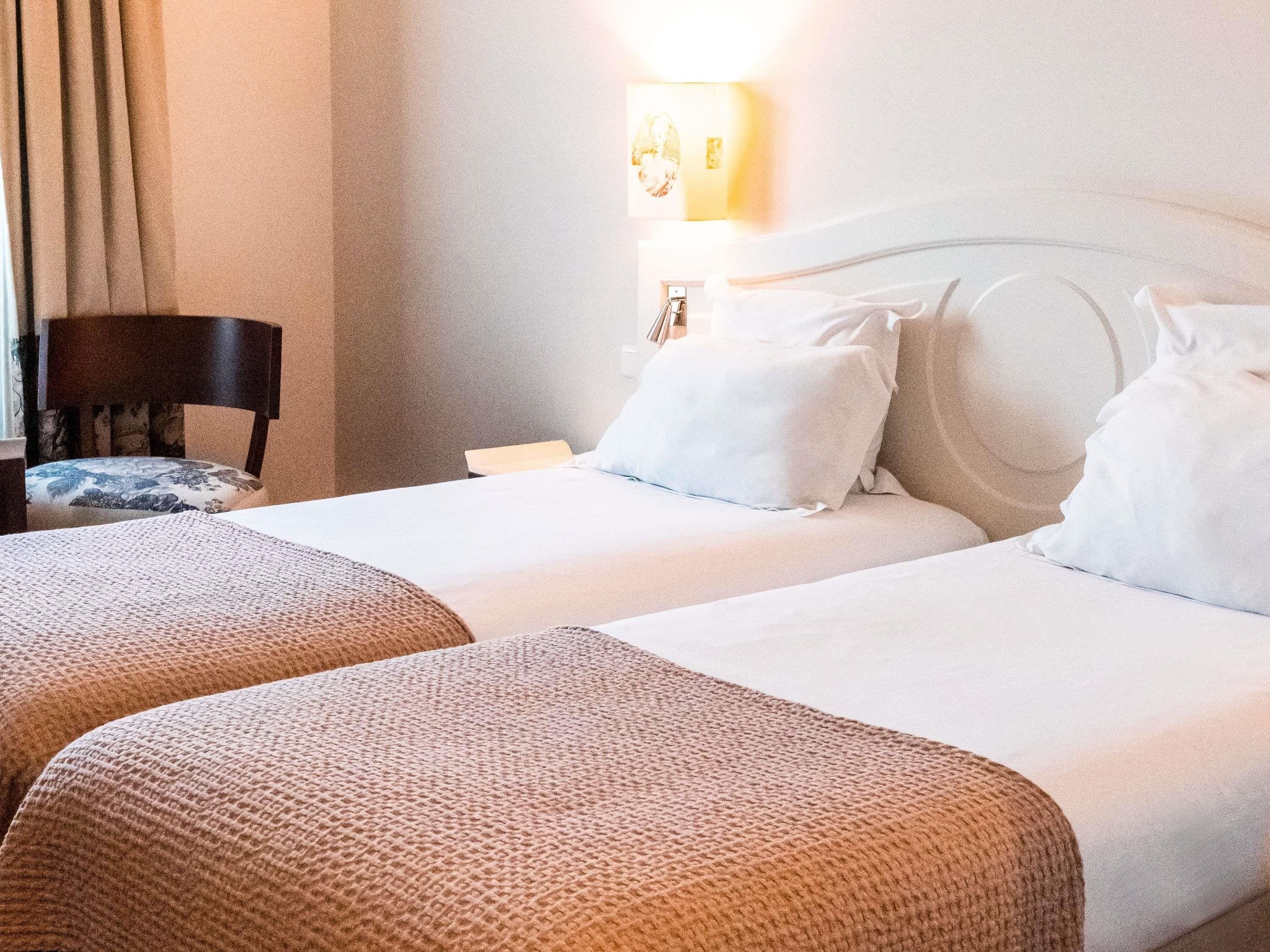 A hotel room with two beds, a chair, a nightstand, and a wall-mounted lamp.