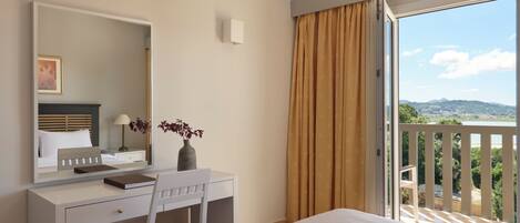 A hotel room with a bed, a desk with a chair, a mirror, a vase with flowers, a window with curtains, and a view of greenery and hills.