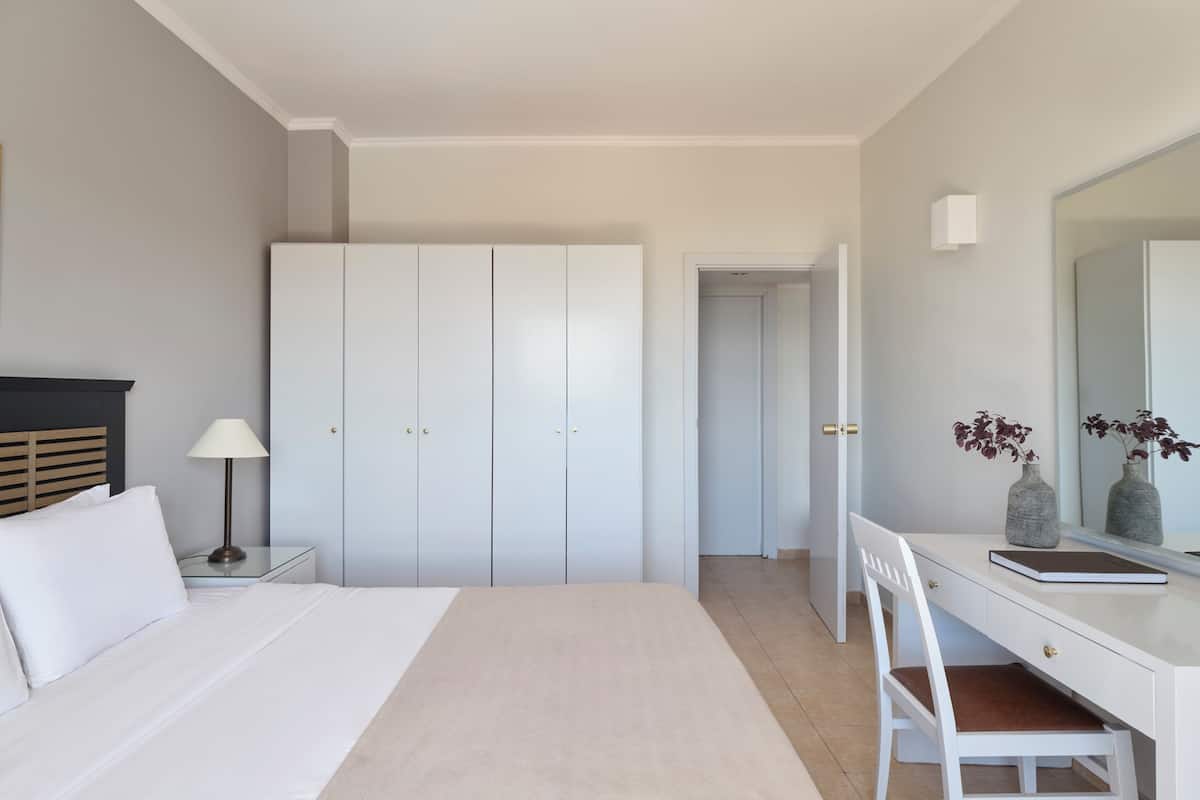 A bedroom with a bed, nightstand, lamp, wardrobe, desk, chair, mirror, and two vases.