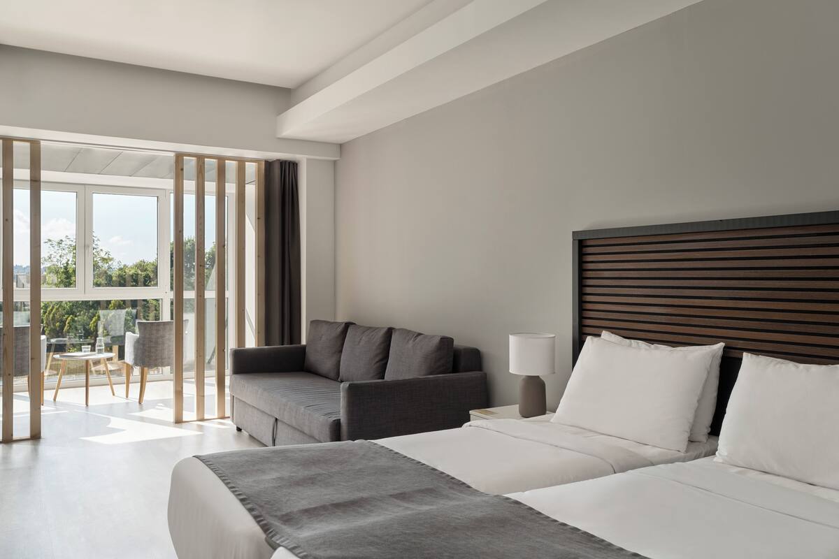 A modern hotel room with a large bed, a sofa, a table, and a view of the outdoors.
