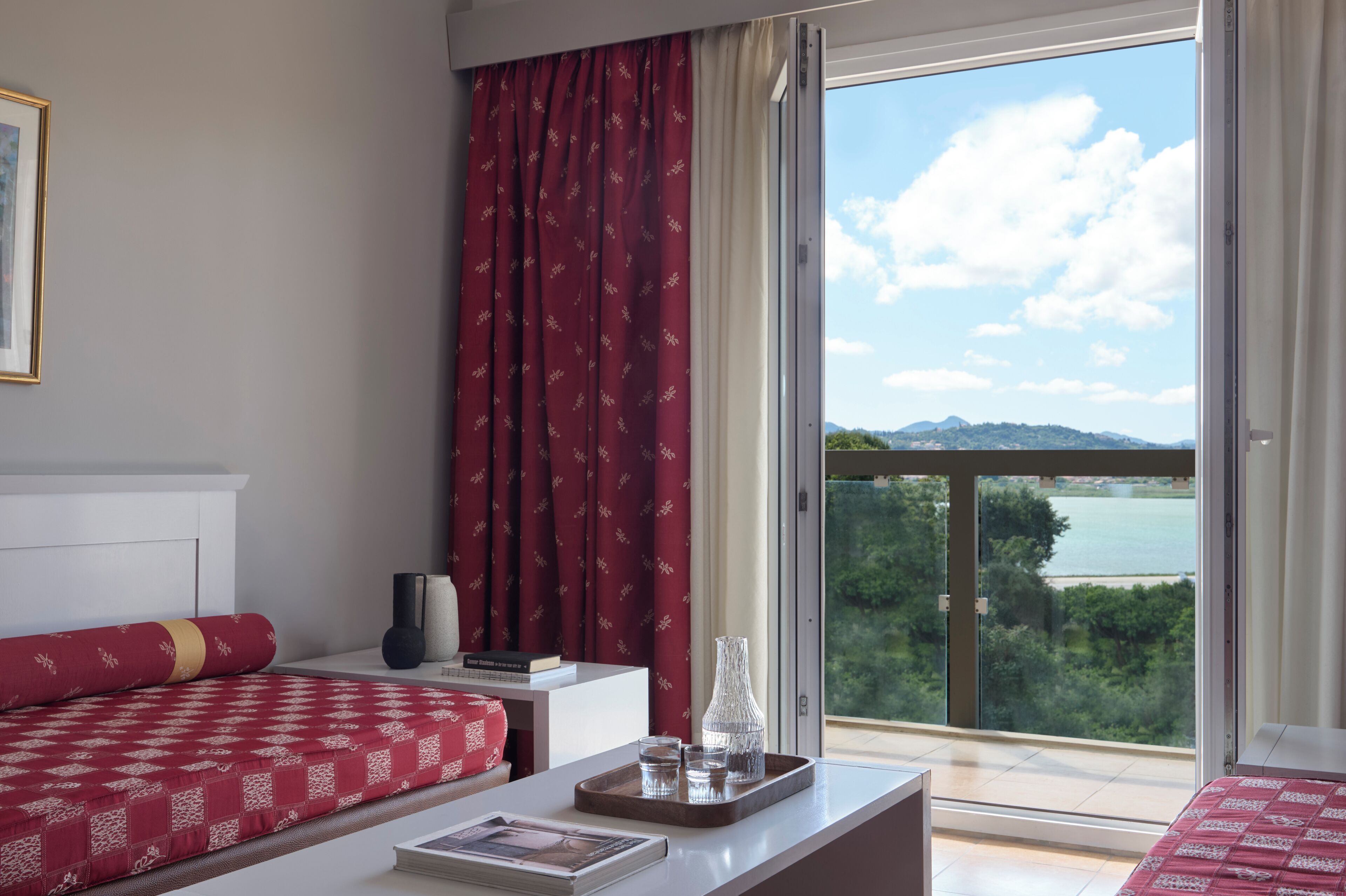 Una habitación de hotel con cama, mesita de noche, ventana con vistas a un cuerpo de agua y vegetación, y balcón.