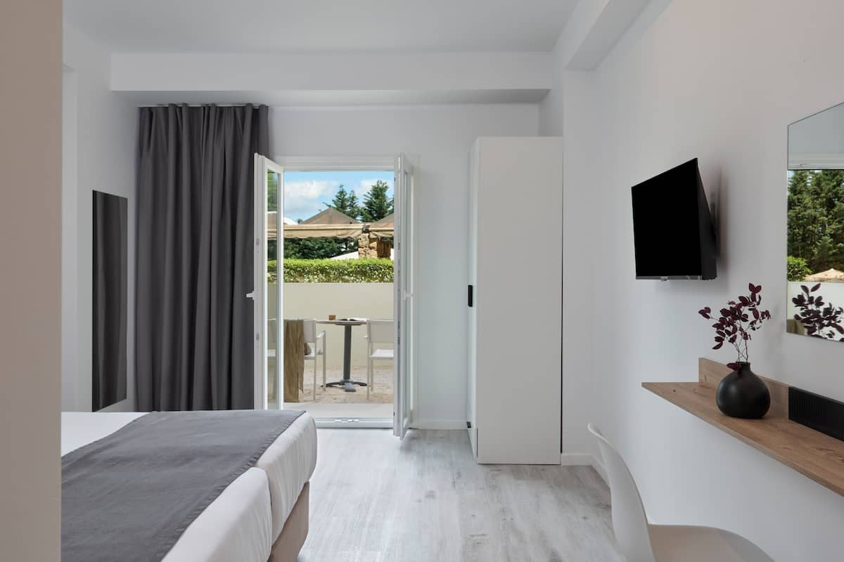 A modern hotel room with a bed, a TV, a wooden side table with a vase, and a view of the outdoor patio.