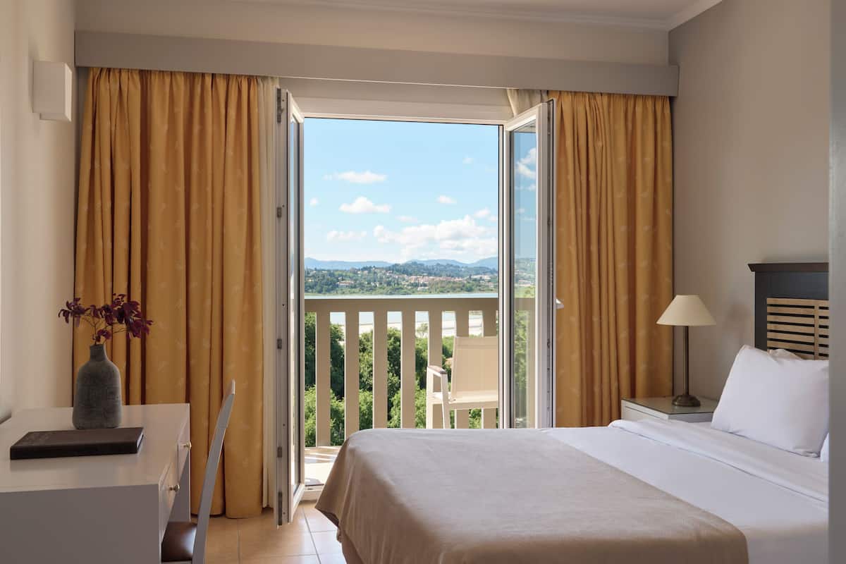 A hotel room with a bed, a desk, a chair, a lamp, and a view of the outdoors.