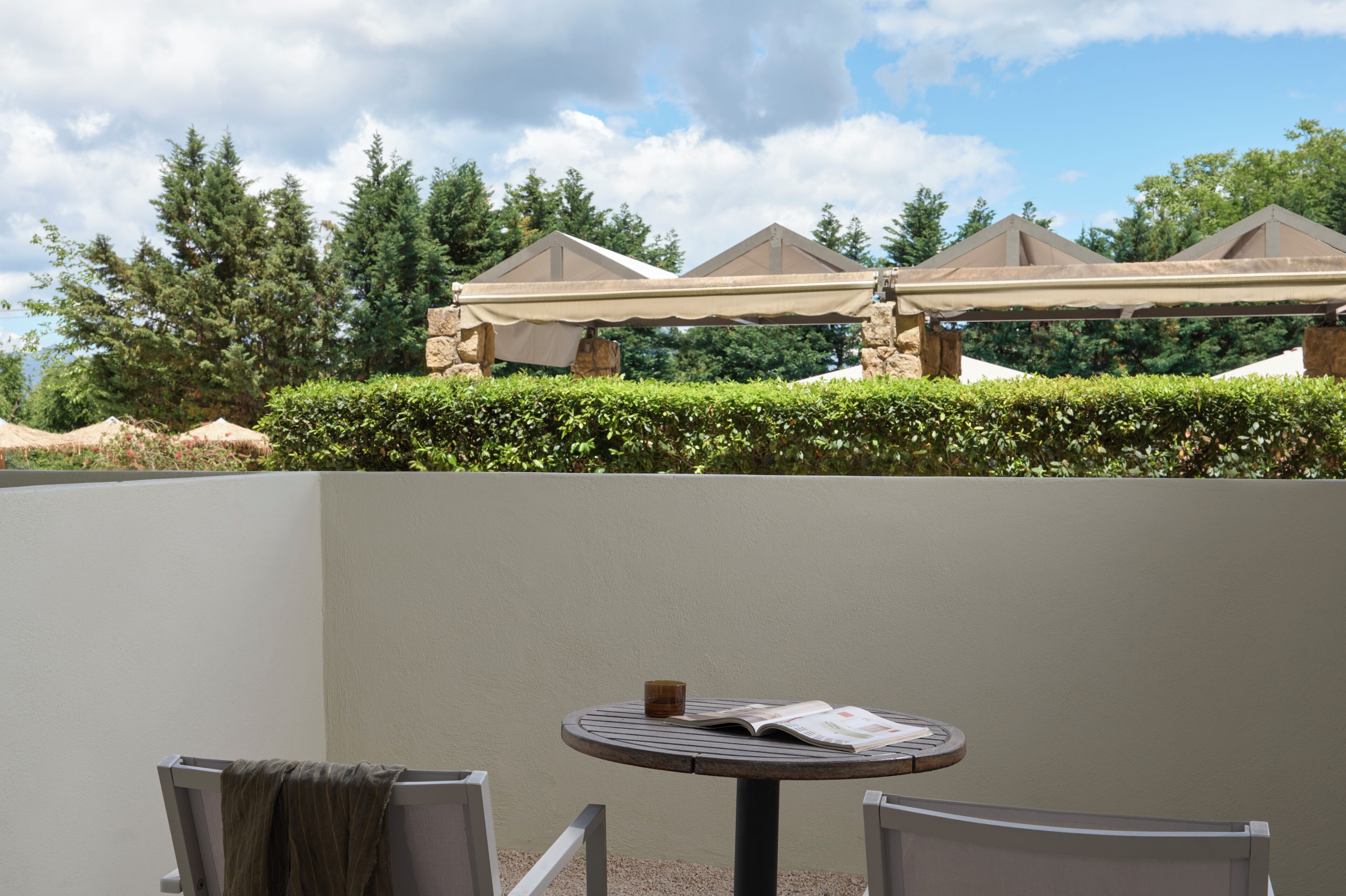 Een terras met een ronde tafel, stoelen en een overdekte zithoek, met uitzicht op bomen en een gebouw op de achtergrond.