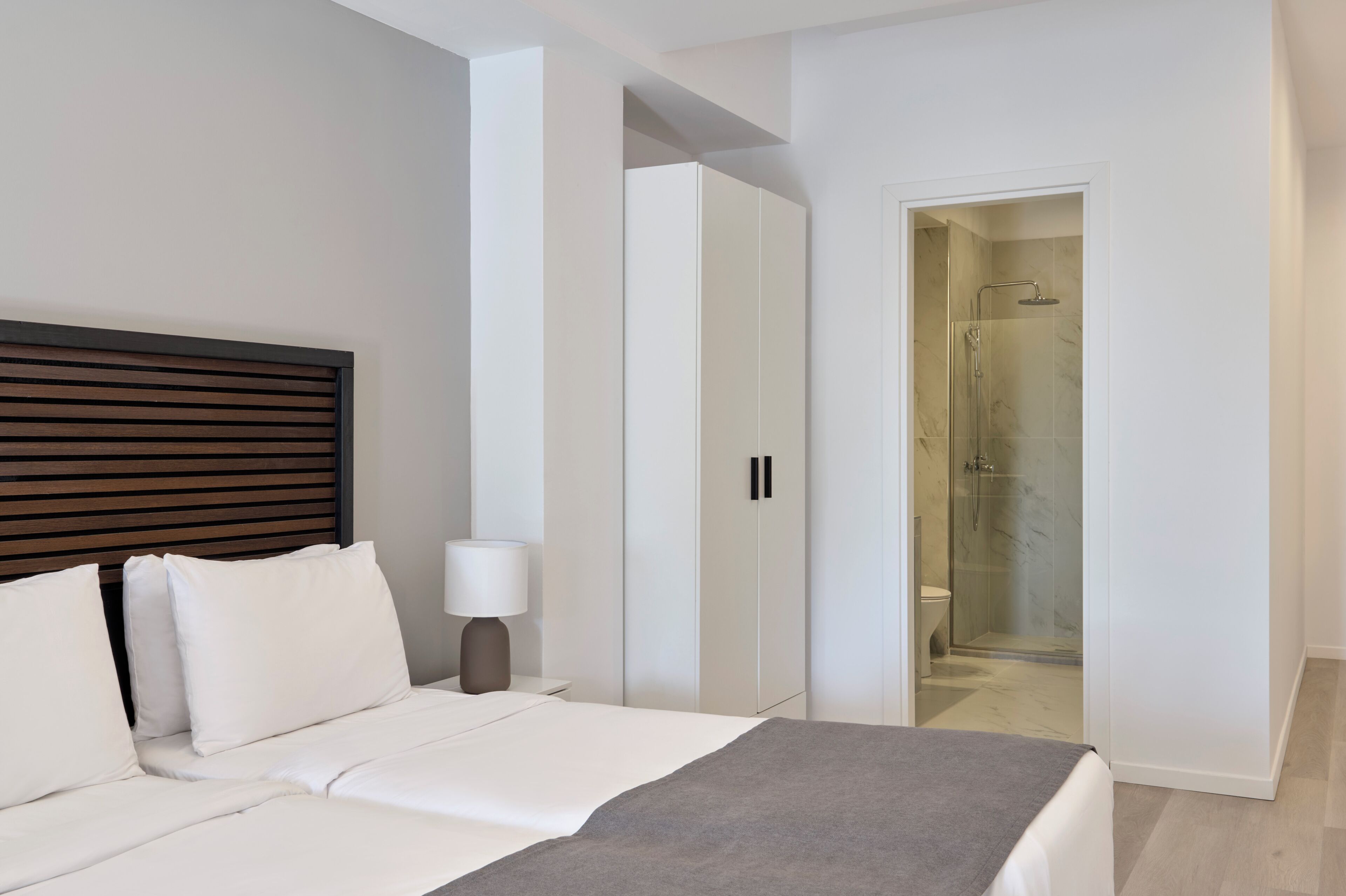 A modern hotel room with a bed, bedside table, lamp, wardrobe, and a shower area.