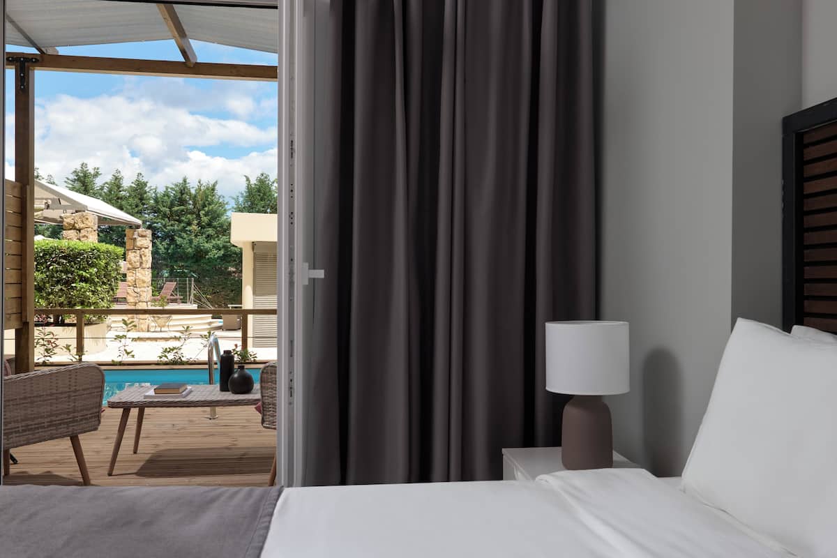 A hotel room with a bed, a nightstand with a lamp, a view of a pool and outdoor area.