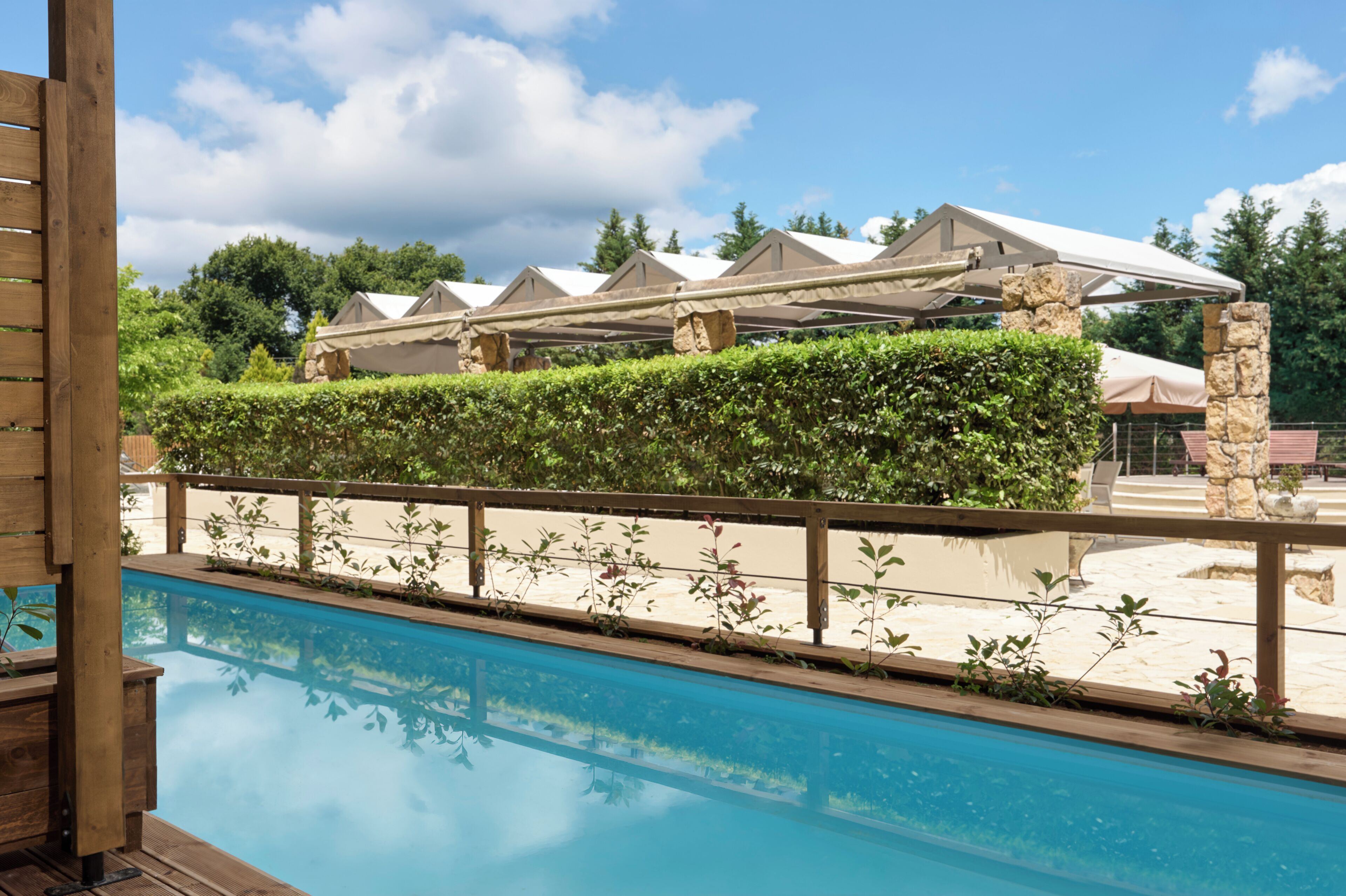 A pool with a wooden deck, a stone wall, and a covered outdoor area with a white roof.