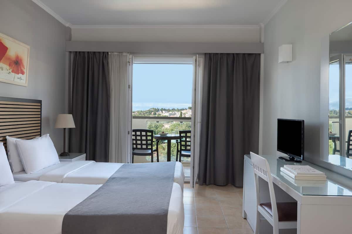 A hotel room with two beds, a desk with a computer, a TV, and a balcony with a view.