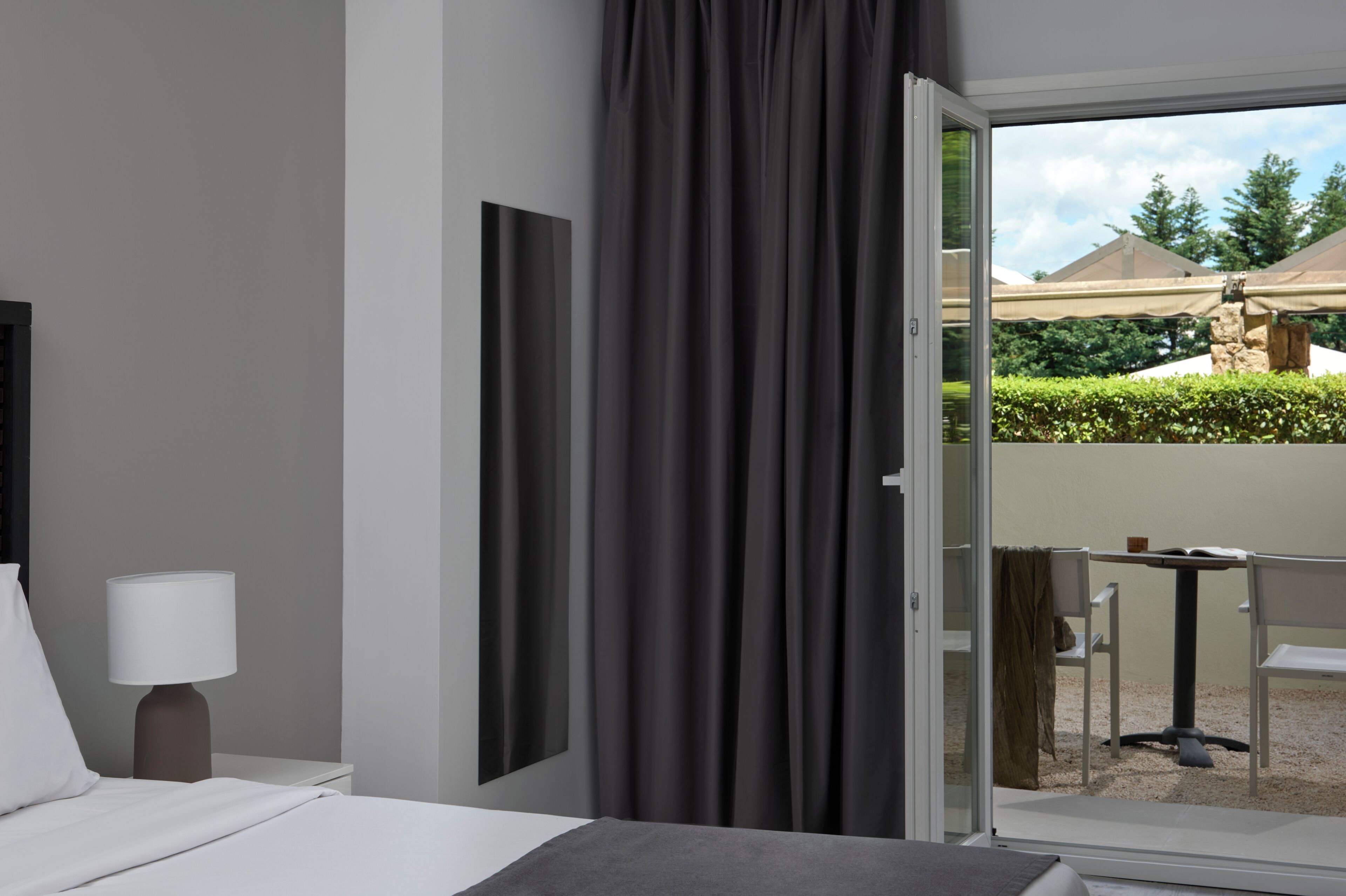A hotel room with a bed, a nightstand with a lamp, a large window with curtains, and a view of an outdoor patio area.