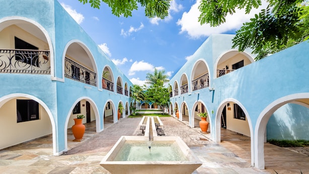 A courtyard with a central fountain, surrounded by blue walls with arched doorways and potted plants. - Hotel Hacienda Inn Aeropuerto (Mérida)