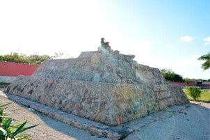 Eine steinerne Pyramidenstruktur mit einer Statue auf der Spitze, umgeben von einer gepflasterten Fläche und einer roten Wand im Hintergrund.