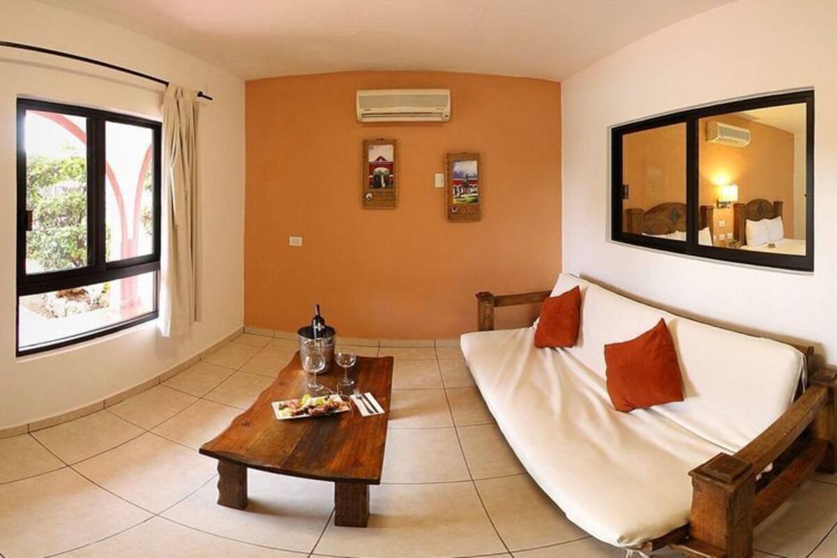 Ein Zimmer mit einer Couch, einem Holztisch mit Flasche und Gläsern, einem Fenster und einer Tür zu einem weiteren Raum.