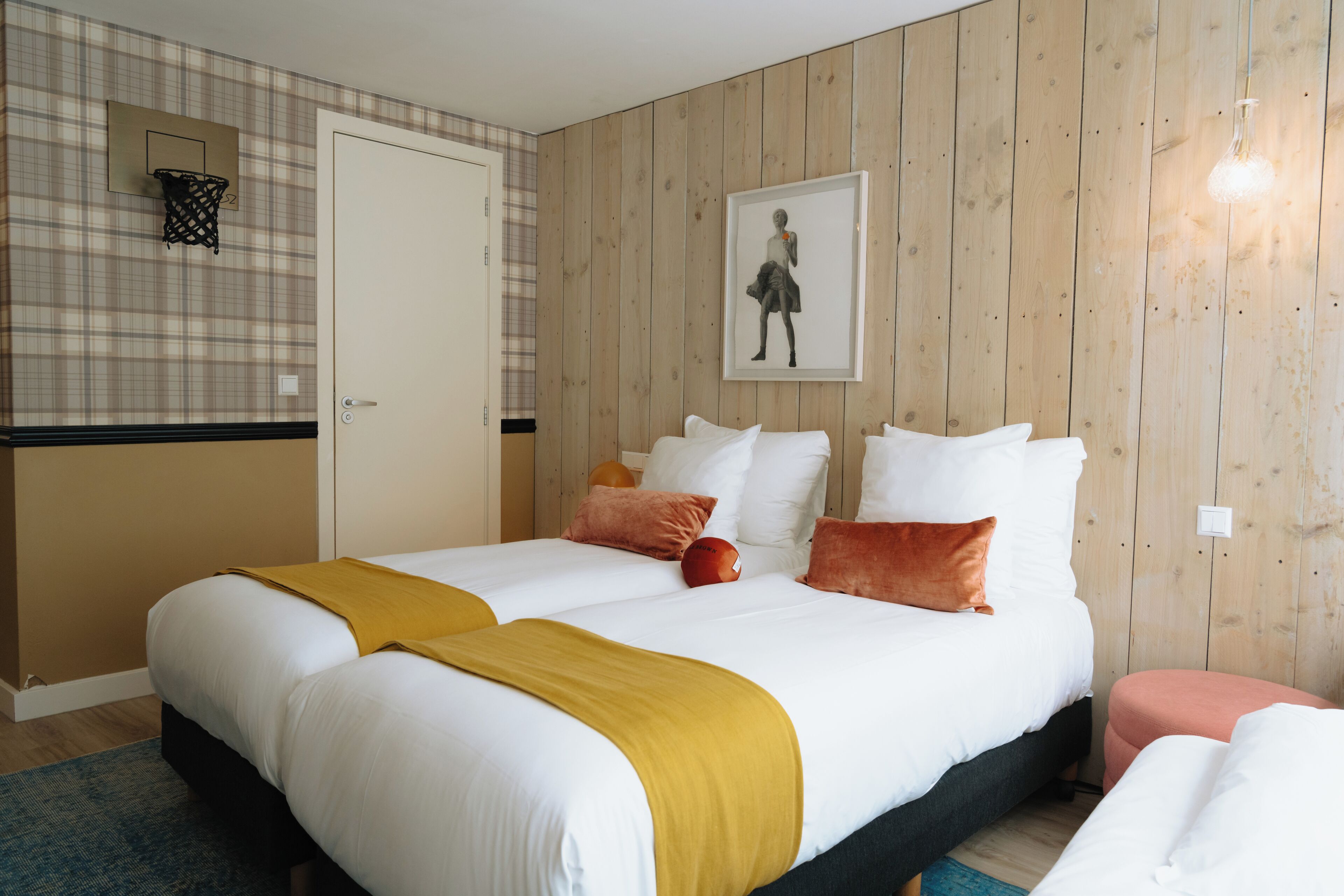A double bed with white and mustard bedding, a wooden wall, a framed picture, and a small basketball hoop.