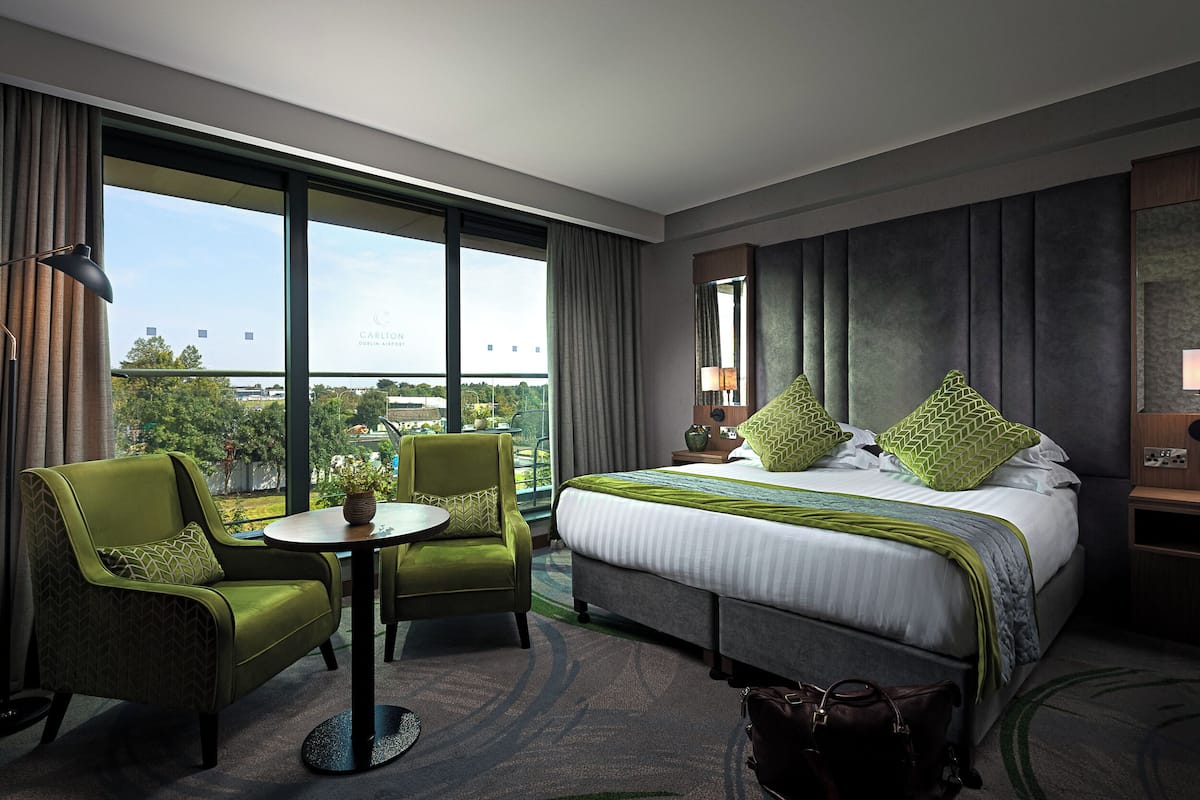 A hotel room with a large bed, two armchairs, a small table, and a view of the airport.