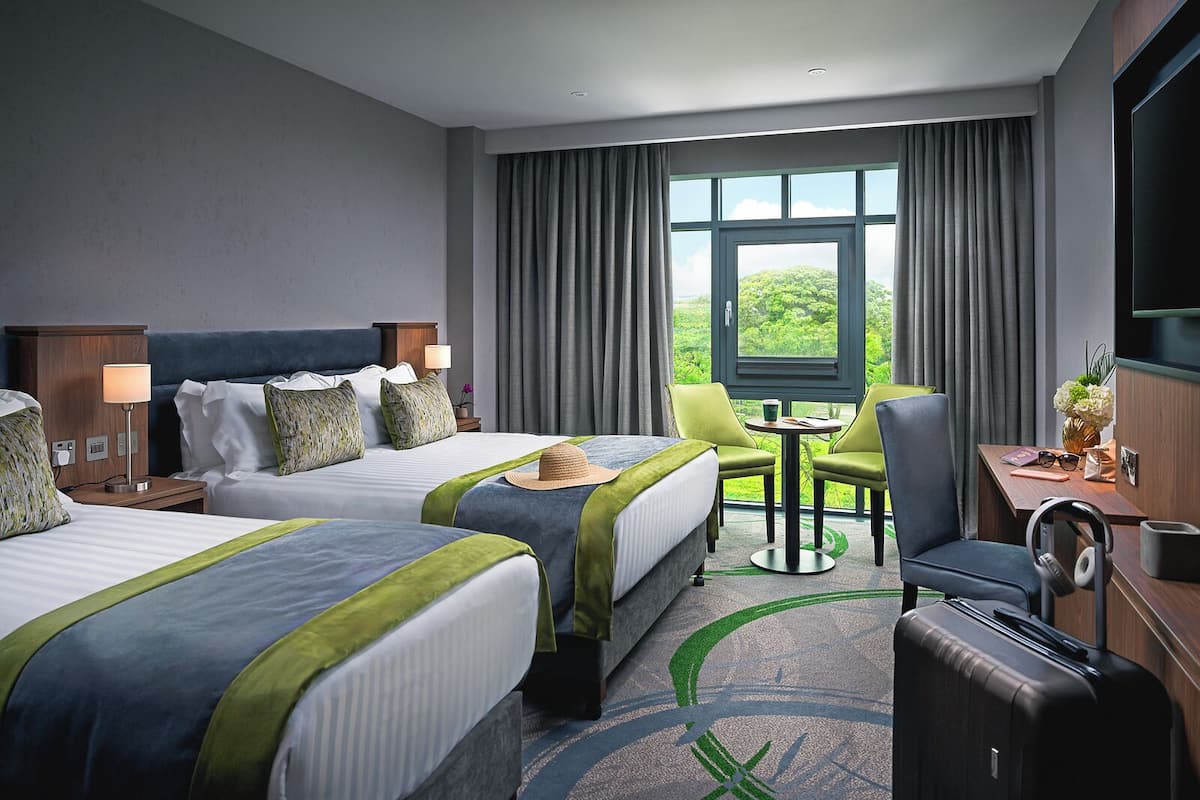 A hotel room with two beds, a desk, a chair, a TV, and a view of greenery through the window.