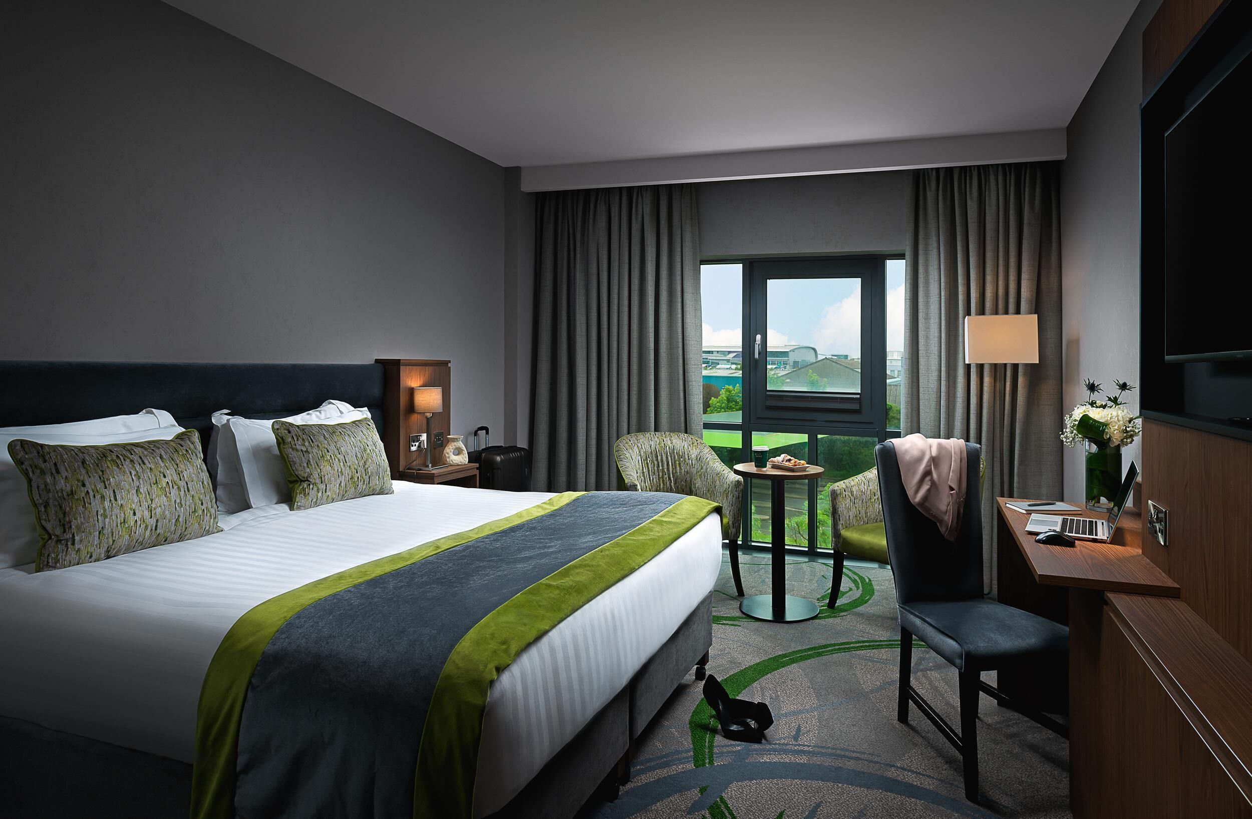 A hotel room with a large bed, a desk with a laptop, a chair, a small table, and a view of the outdoors.