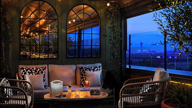 Un rincón lounge acogedor con sofá, sillas, una mesa y ventanas arqueadas amplias que dan a vista del aeropuerto.