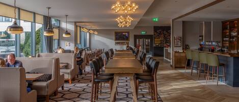 A modern restaurant with a long wooden table, comfortable seating, and a bar area.