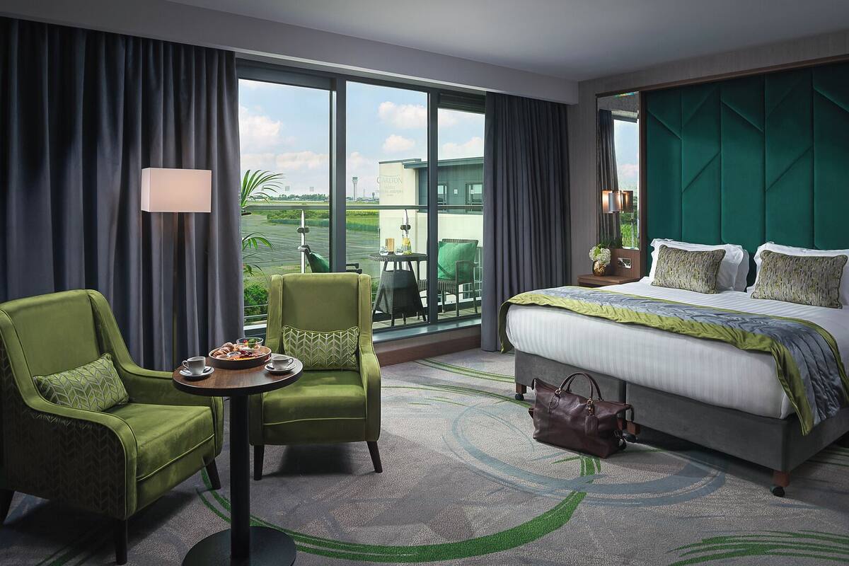 A hotel room with a large bed, a green armchair, a small table with breakfast, and a view of a green landscape through large windows.