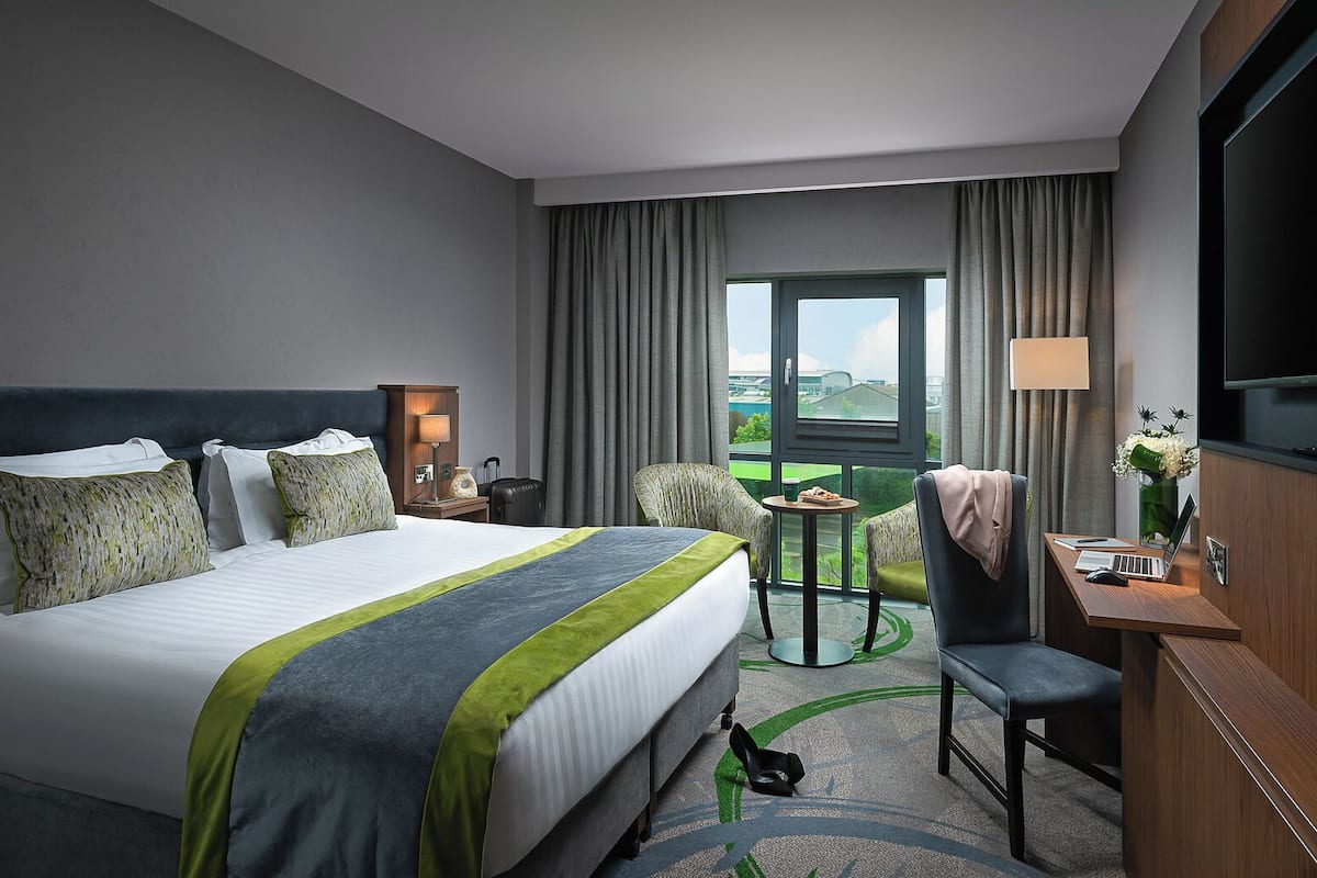 A hotel room with a large bed, a desk with a laptop, a chair, a small table with a drink, and a view of the outdoors.