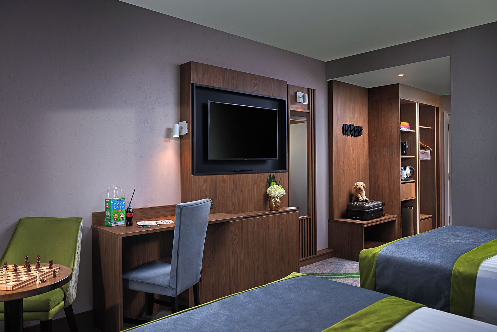 A hotel room with a TV mounted on the wall, a desk with a chair, a small table with chess, two beds with green and blue bedding, and a wardrobe with a mirror and shelves.