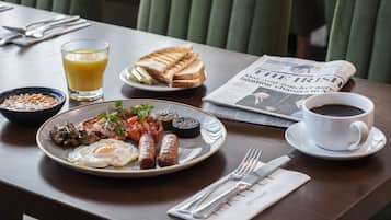 A breakfast table with a plate of eggs, sausages, mushrooms, and toast, a glass of orange juice, a cup of coffee, and a newspaper.