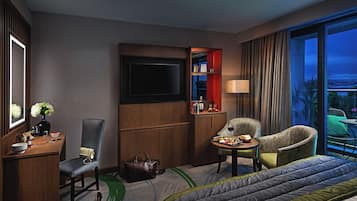 Habitación de hotel con cama, escritorio con silla, televisor, minibar y vistas al exterior.