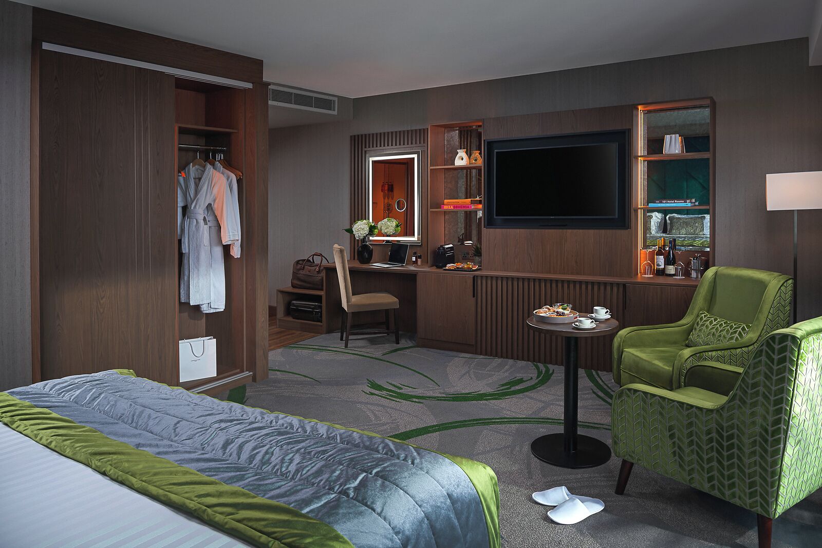 Quarto de hotel com cama, escrivaninha com cadeira, televisão, guarda-roupa e área de estar com poltrona.