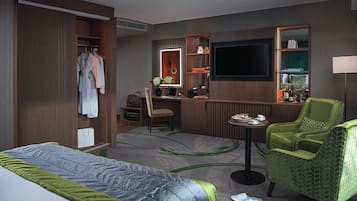 Quarto de hotel com cama, escrivaninha com cadeira, televisão, guarda-roupa e área de estar com poltrona.