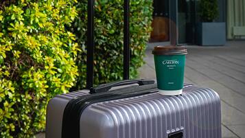 A silver suitcase with a green coffee cup on top, placed in front of a building with a 'Carlton' sign.