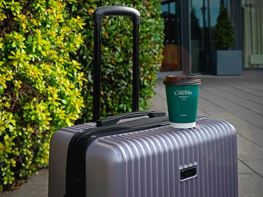 A silver suitcase with a green coffee cup on top, placed in front of a building with a 'Carlton' sign.