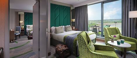 A hotel room with a bed, a green armchair, a small table with a plant and two cups, and a view of the outdoors through large windows.