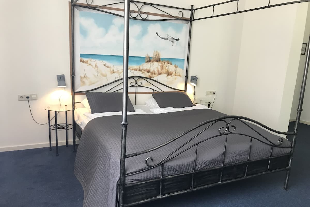 Una cama con dosel, colcha gris, dos lámparas de noche y un cuadro con una playa en la pared.