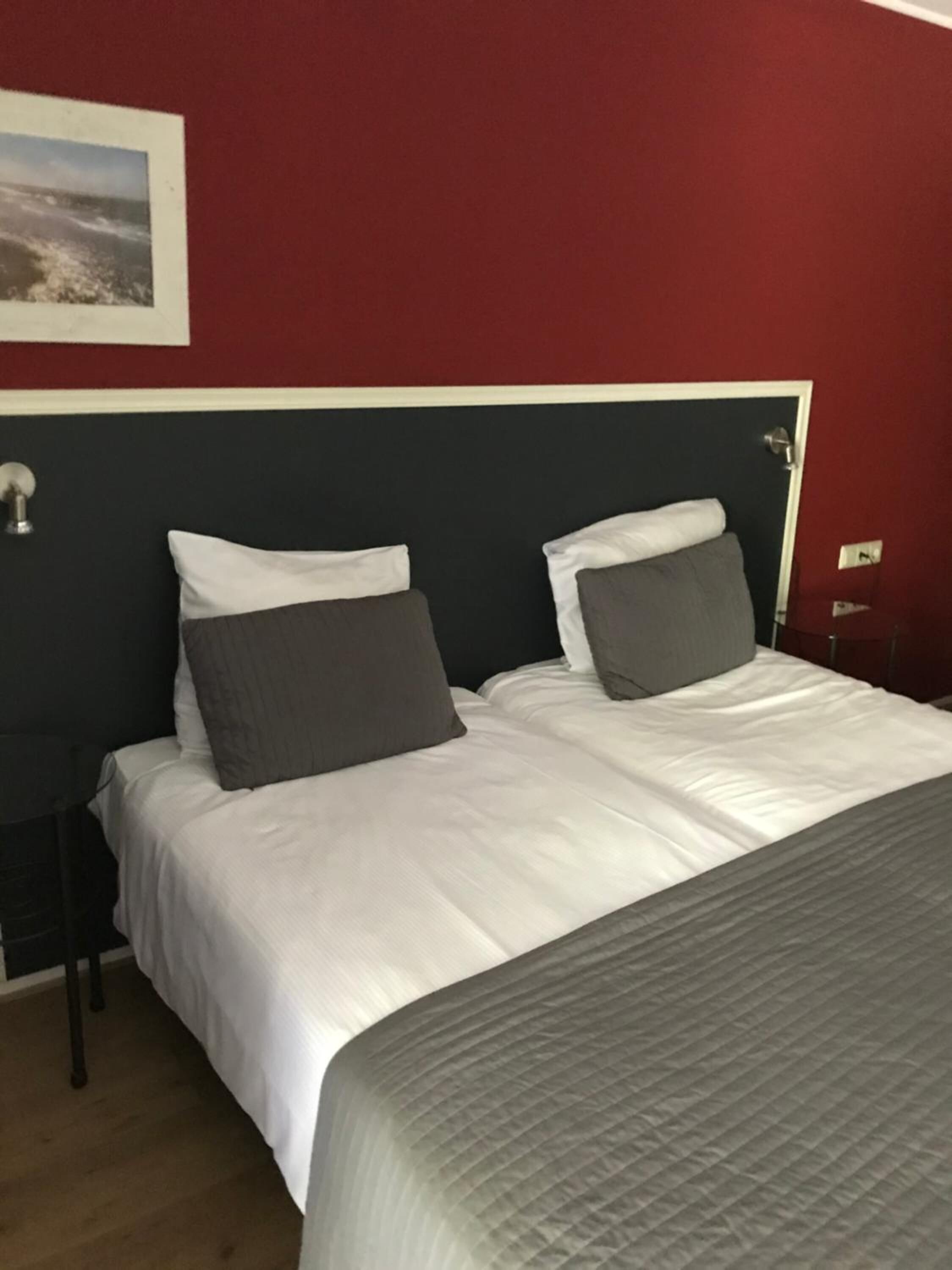 Een tweepersoonsbed met wit en grijs beddengoed, een donker hoofdbord en een rode muur met daarboven een ingelijste foto.