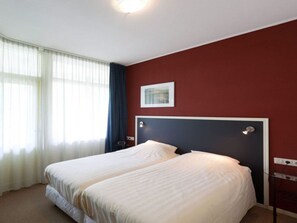A hotel room with two beds, a red accent wall, a framed picture, and a window with sheer curtains.