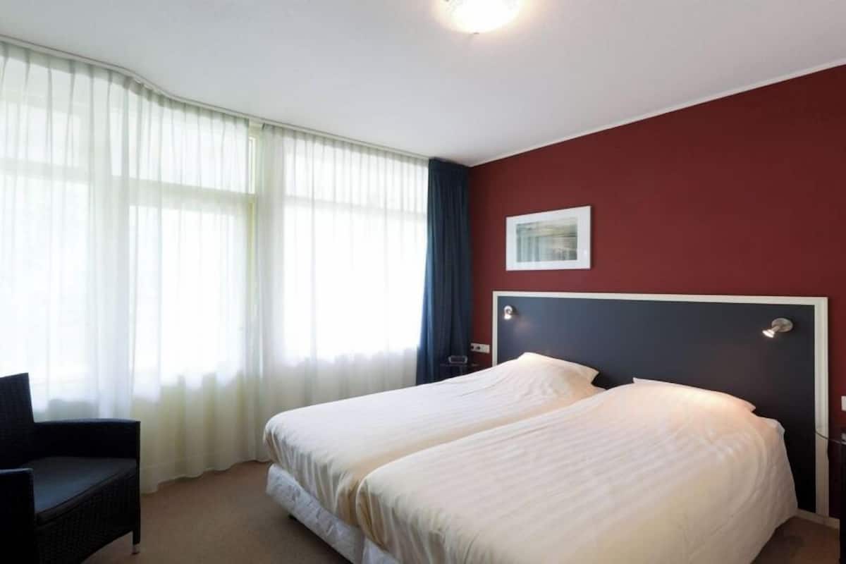 Una habitación de hotel con cama, una silla, ventana con cortinas y un cuadro en la pared.