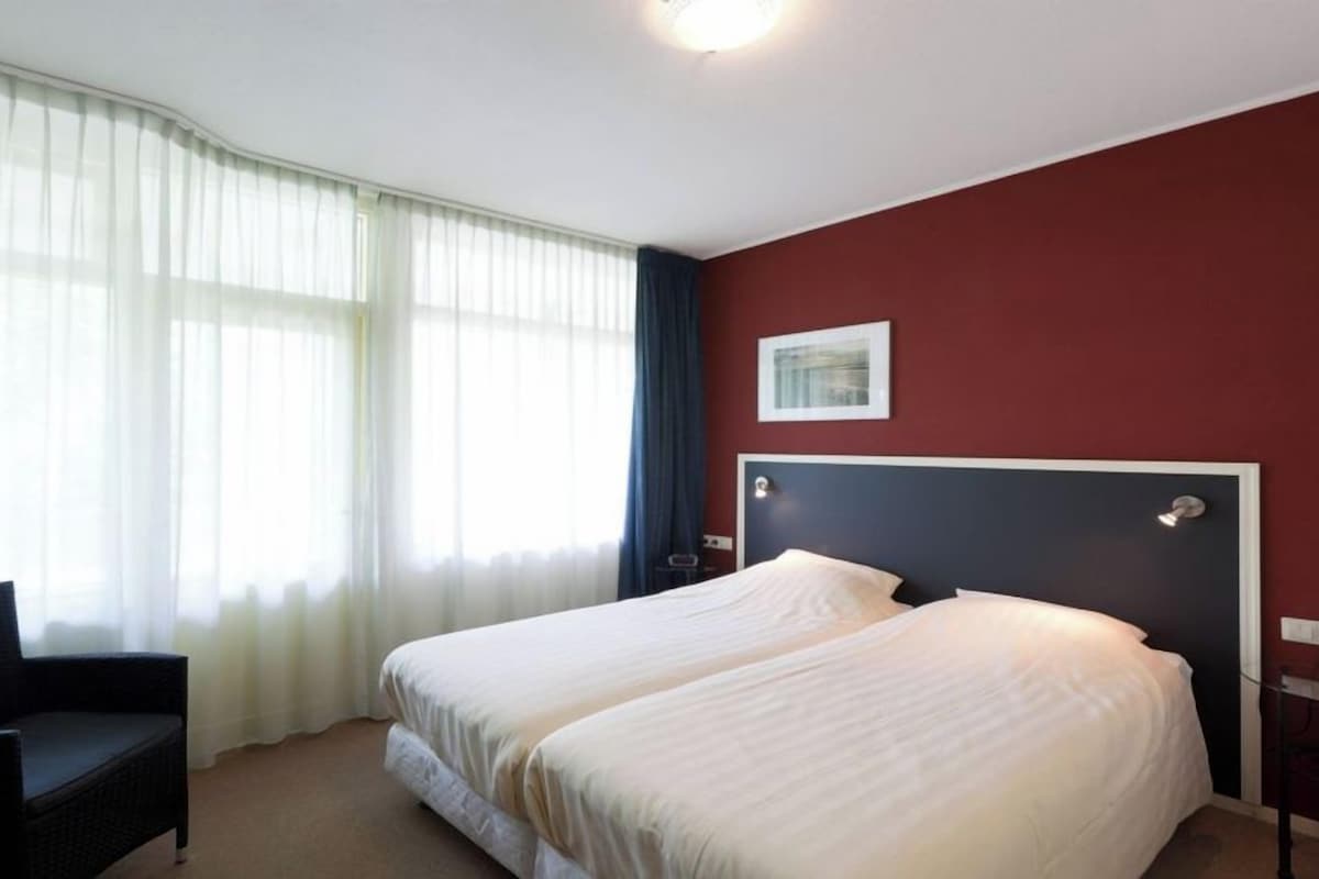 A hotel room with a bed, a chair, a window with curtains, and a wall-mounted picture.