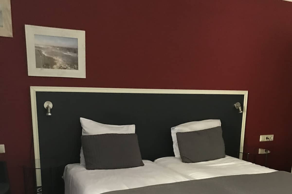 Una cama bien hecha con colcha gris, cabecera oscura y pared roja con un cuadro enmarcado encima.