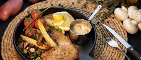 Un plato con bistec, papas fritas y vegetales, acompañado de una bebida y cubiertos sobre un posavasos tejido.