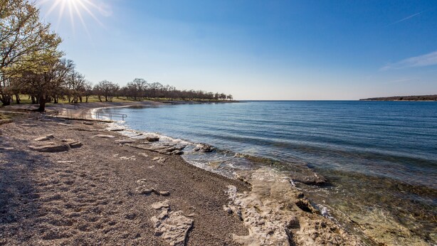 A beach with clear water, pebbles, and trees under a bright sun. - Apartment House Volta (Porec)