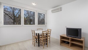 A room with a dining table and chairs, a television on a wooden stand, and large windows with a view of trees. - Apartment House Volta (Porec)