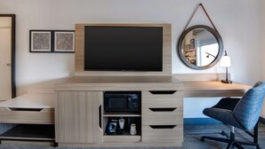 A modern hotel room with a flat-screen TV mounted on a wooden console, a microwave, and a coffee maker. There is a round mirror on the wall and a chair with wheels.