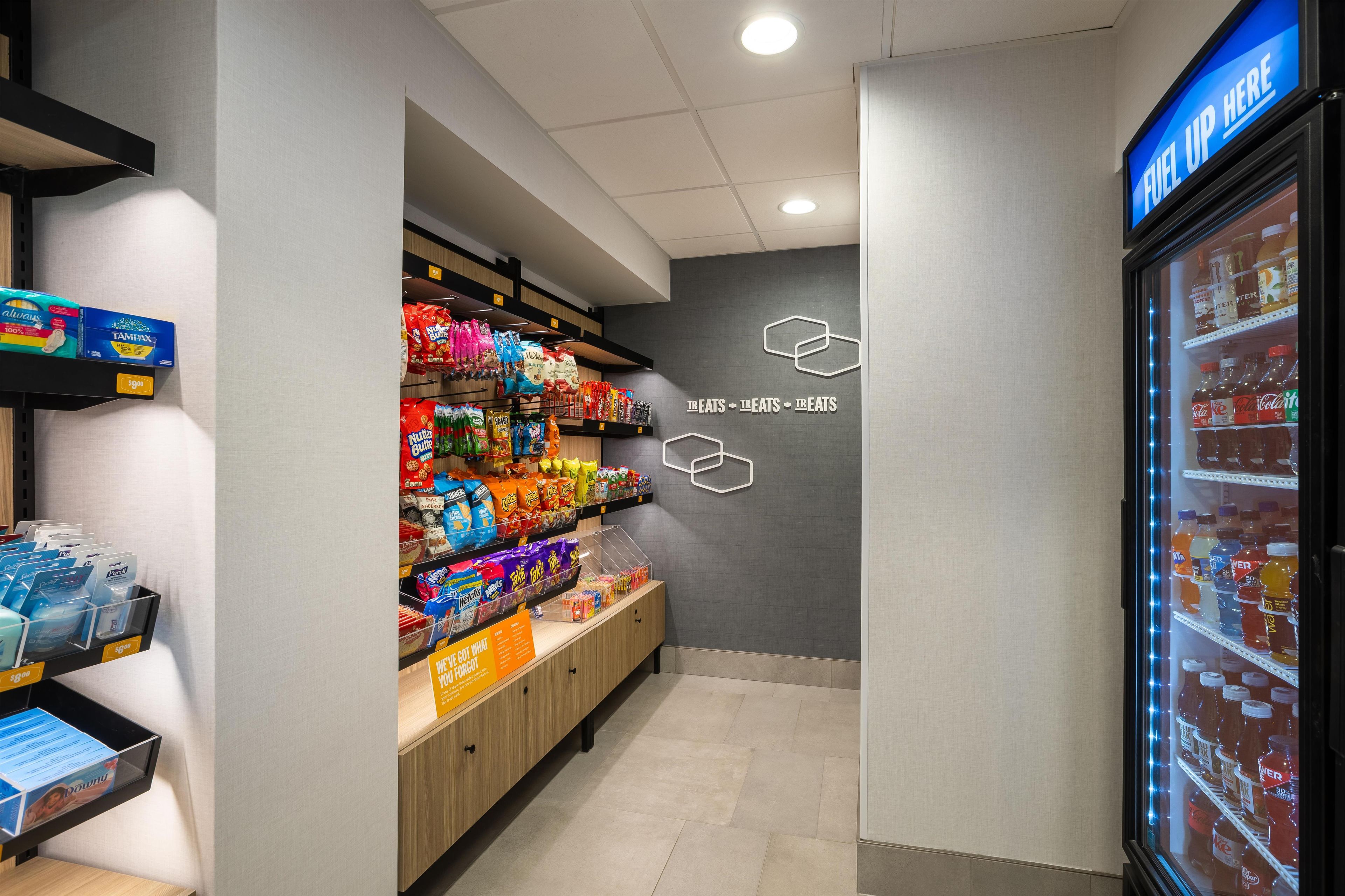 A store aisle with shelves stocked with various snacks and beverages, a refrigerated section with drinks, and a sign indicating a fuel station.