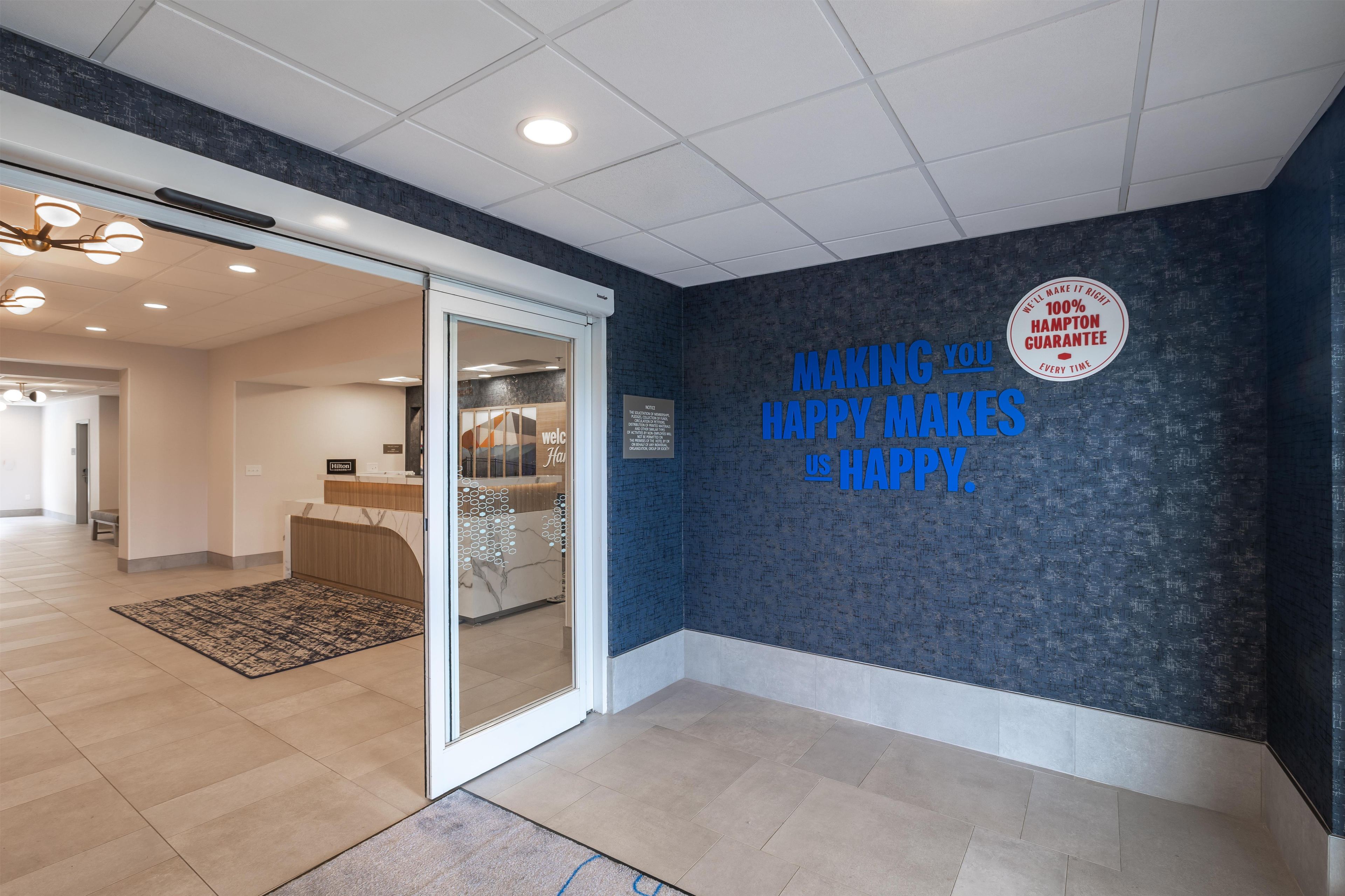 A hotel reception area with a dark textured wall, a glass door entrance, and a sign stating 'Making you happy makes us happy.'