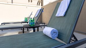 A row of sun loungers with towels, a small table with bottles, and a building in the background.