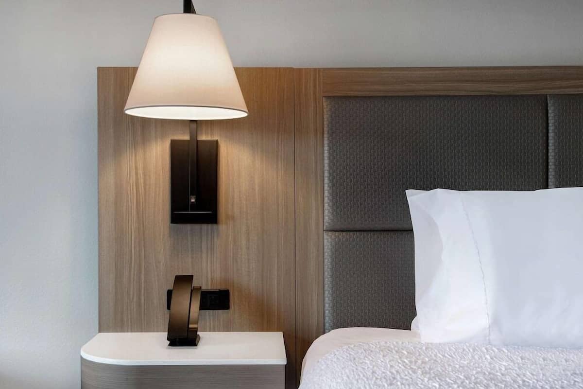 Una habitación de hotel con una cama, una mesita de noche, una lámpara y un televisor montado en la pared.