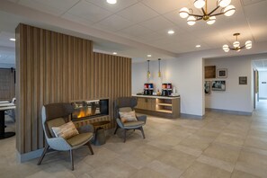 A modern living area with a fireplace, two armchairs, a coffee station, and a dining table.